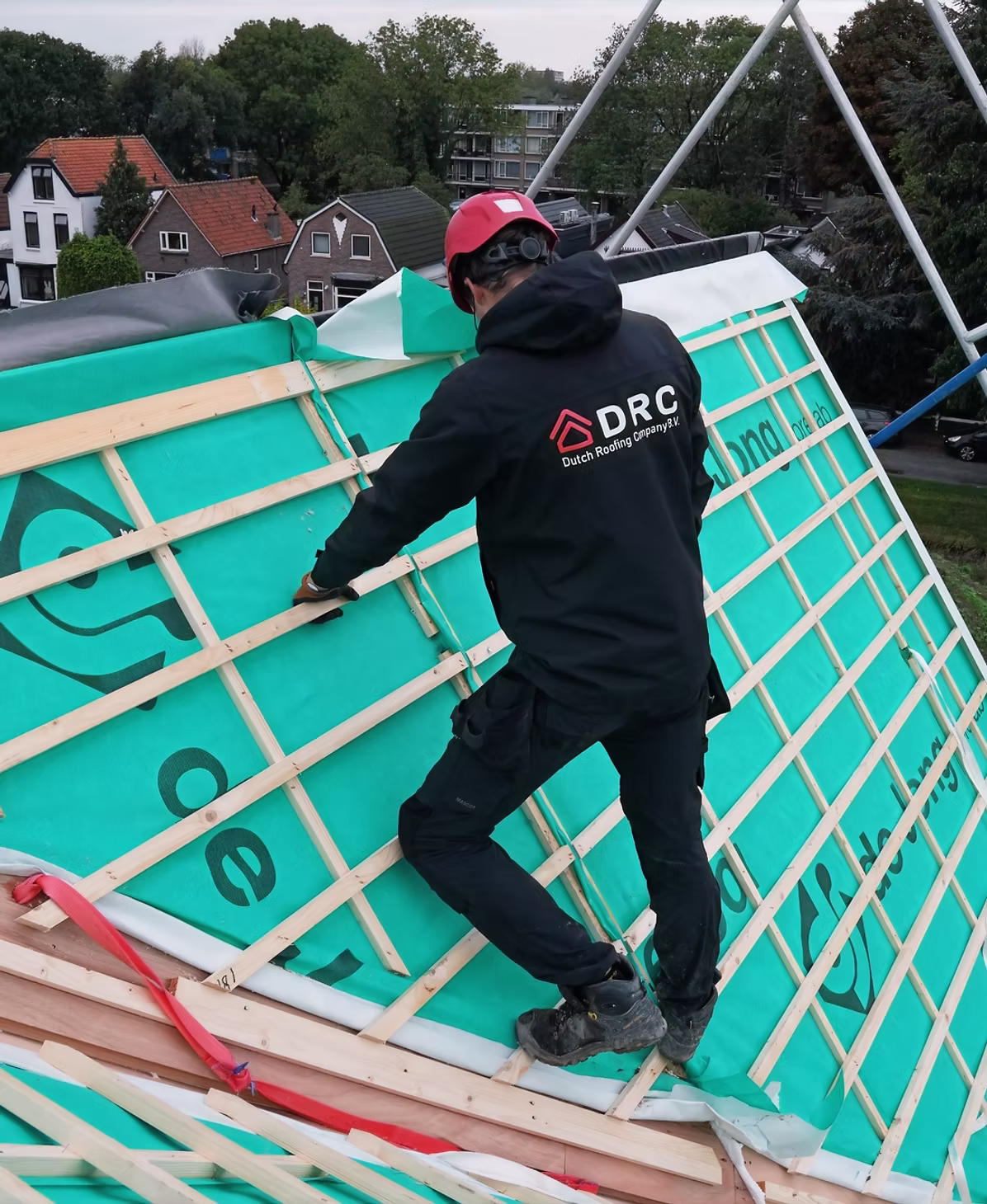 Een man met een rode helm en een zwarte jas met het logo DRC plaatst een groene waterdichte folie op een hellend dak van een huis, met houten latten als dakconstructie.