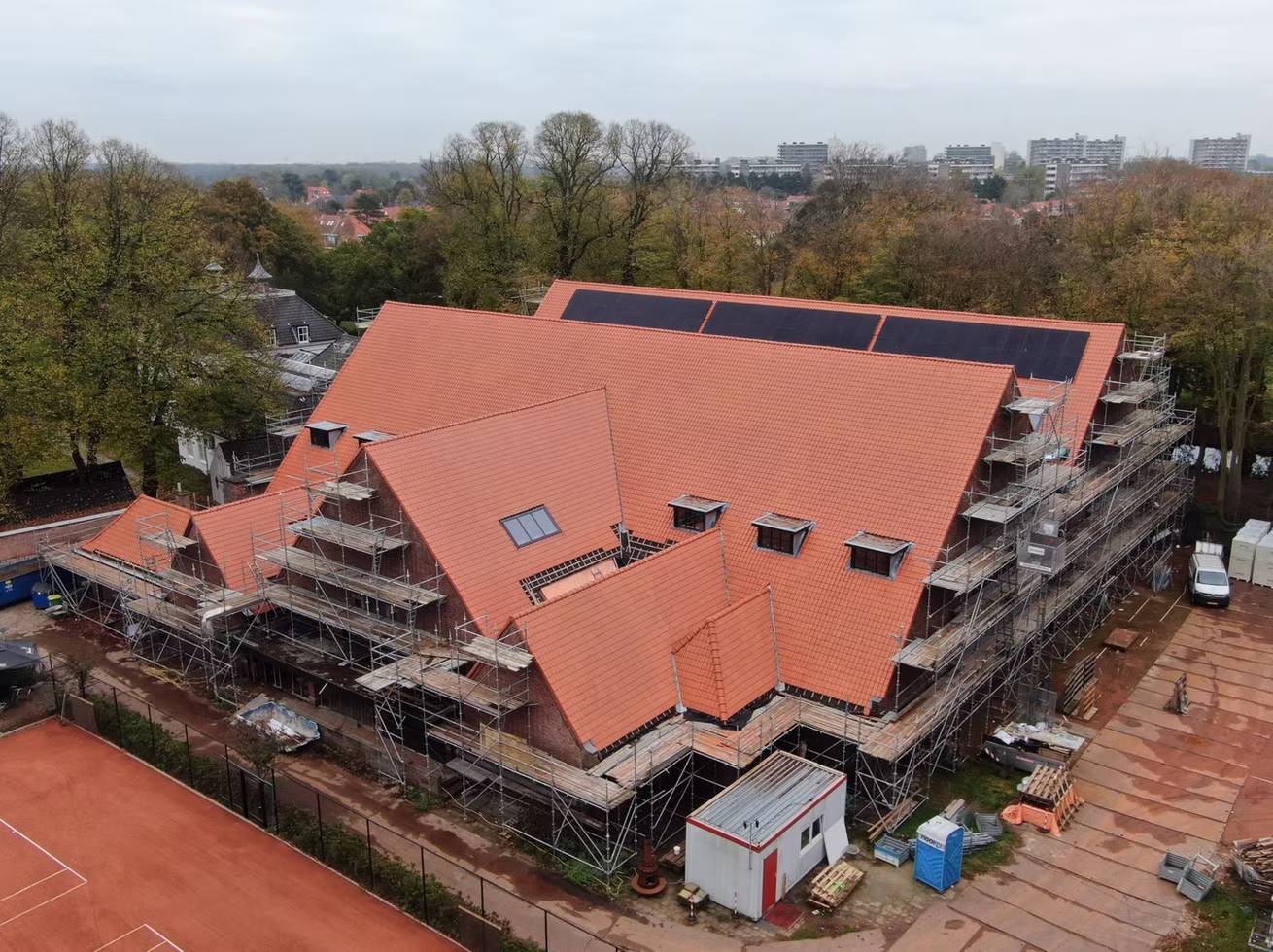 Groot gebouw met een rood dak en zonnepanelen, omringd door steigers en een tennisbaan.