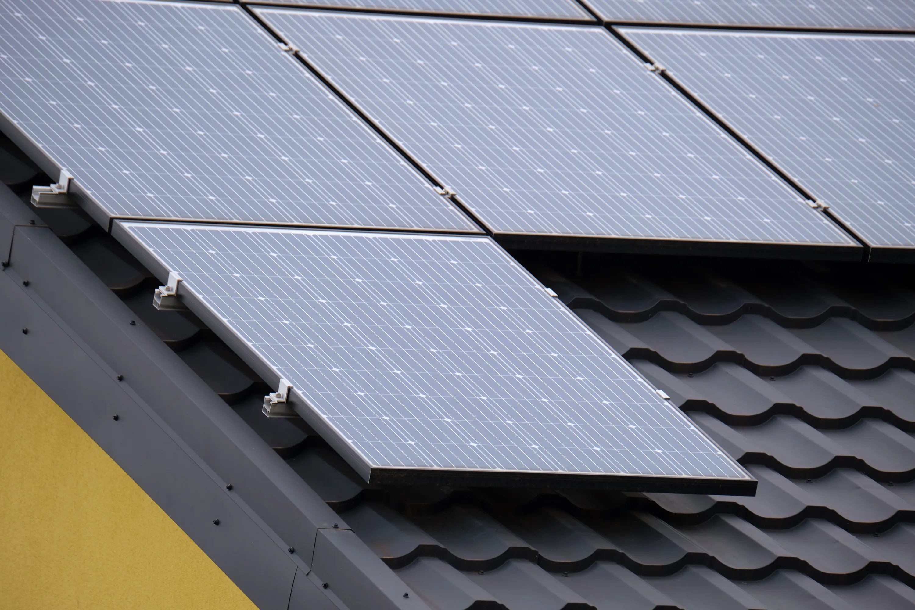 Aurinkopaneelit asennettuna tummalle tiilikatolle, keltainen seinä näkyy vasemmalla. Paneelit ovat harmaat ja niissä on valkoisia raitoja.
