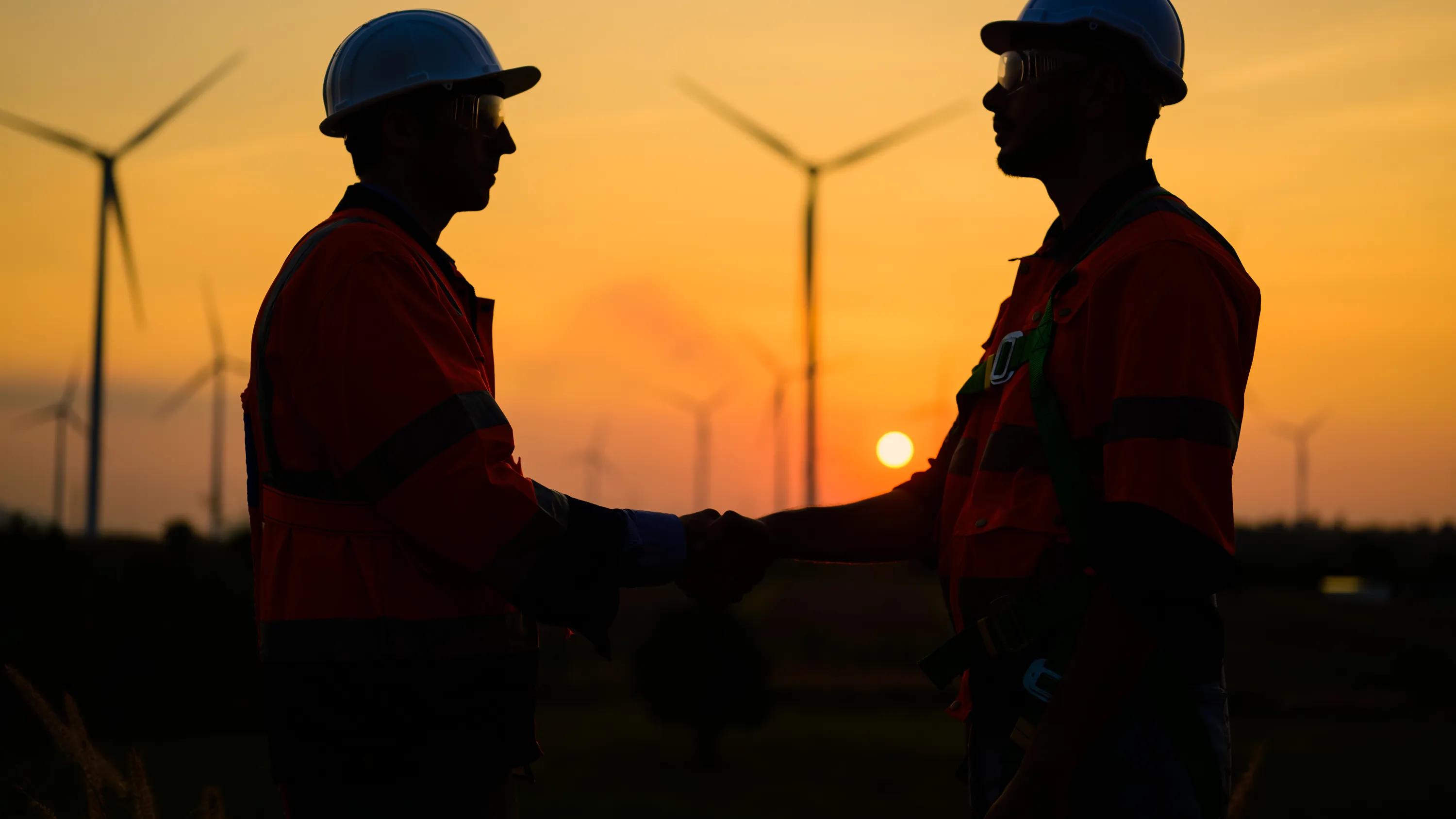 Siluettikuvassa kaksi insinööriä kättelee tuulivoimaloiden taustalla auringonlaskun aikaan. Heillä on yllään suojakypärät ja huomiovaatteet.