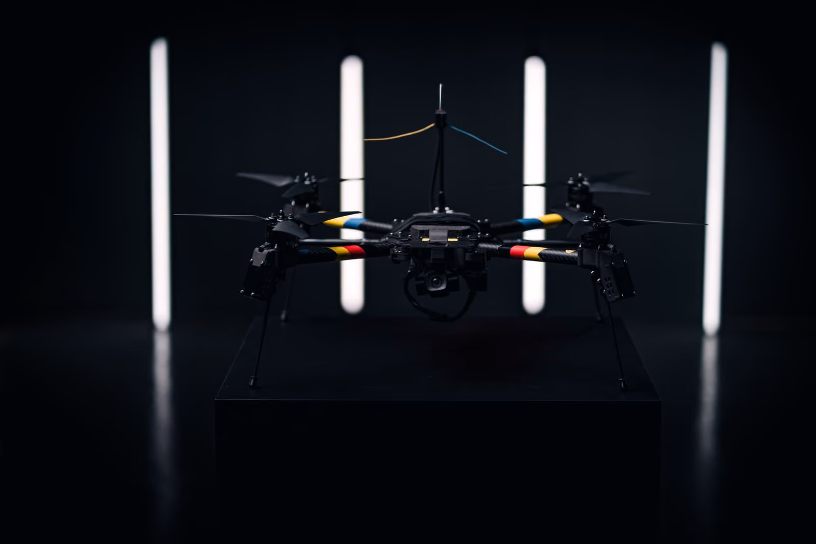 Black quadcopter drone from Quantum Frontline Industries with colored bands on arms displayed in dark room under vertical lights.