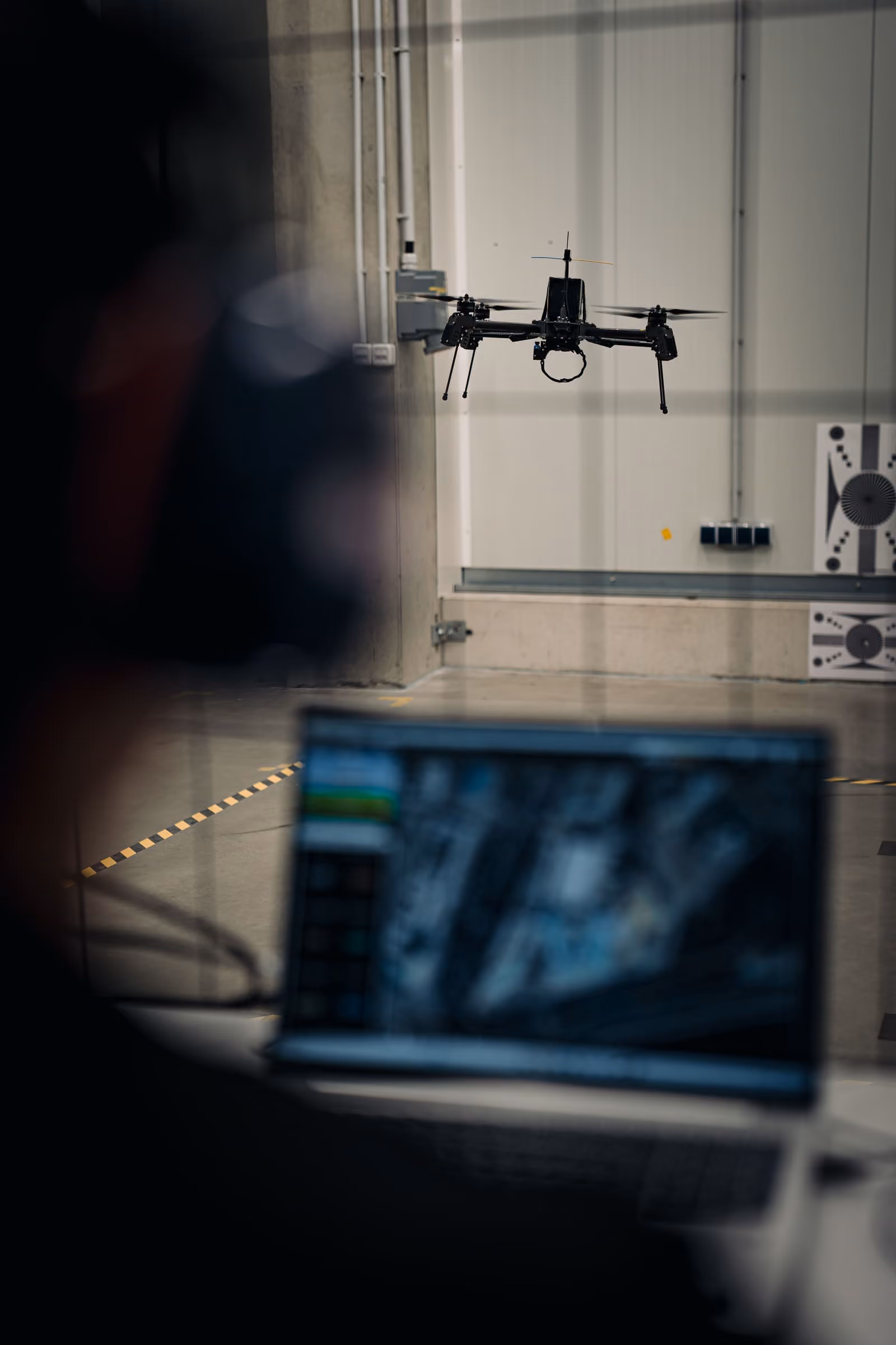 Black Quantum Frontline Industries drone hovering indoors with a blurred laptop and person controlling it in the foreground.