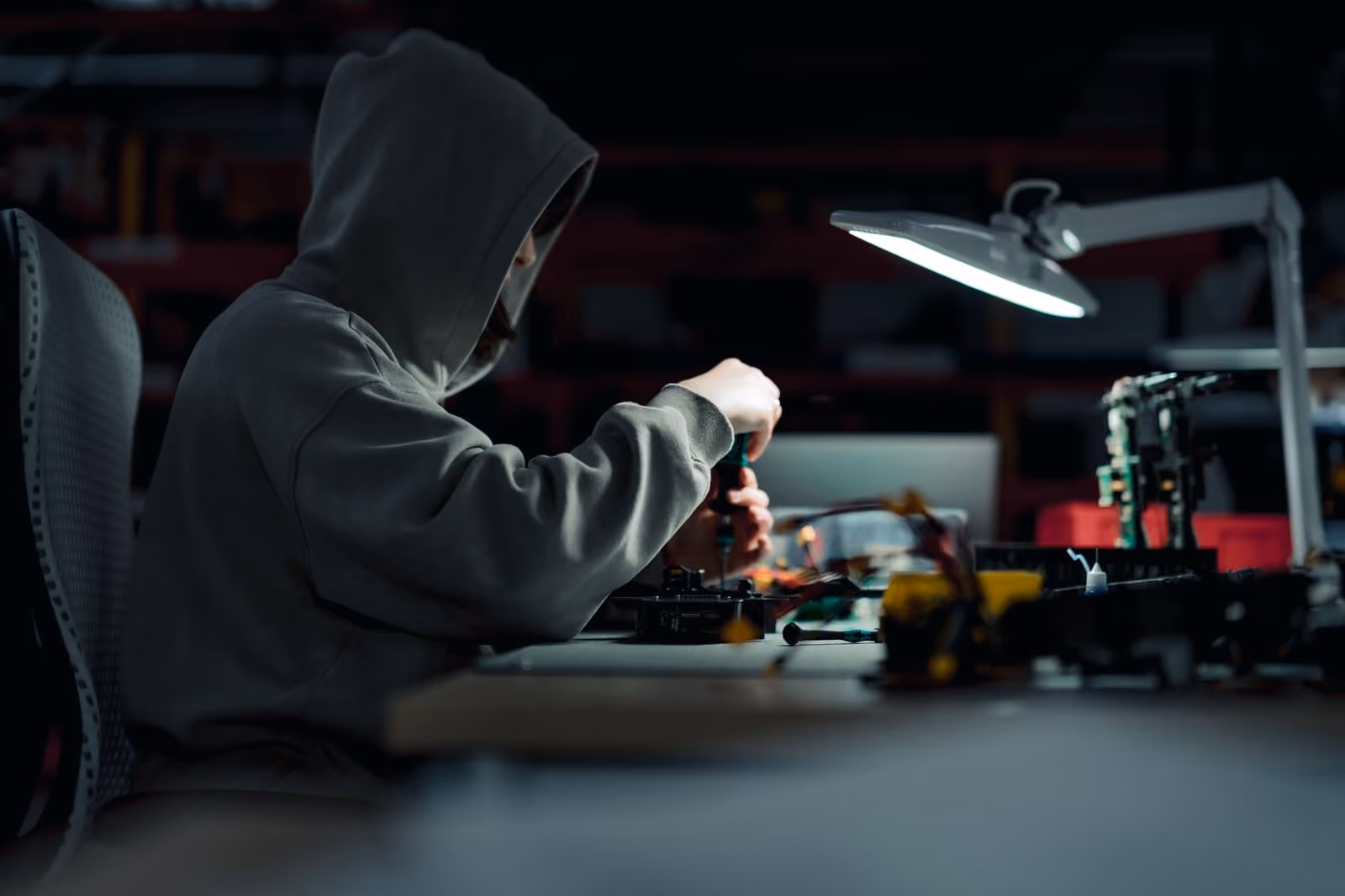 Person in hoodie assembling or repairing a drone under a desk lamp in a dim workspace.
