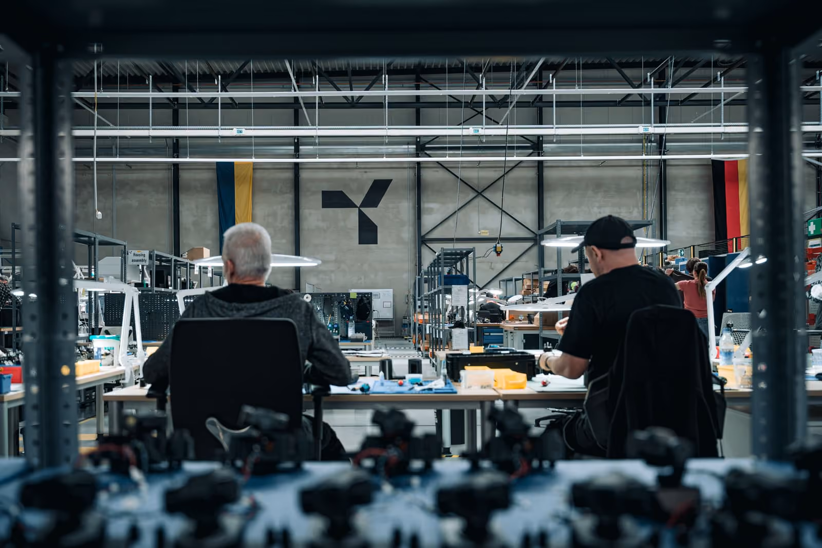Two workers seated at desks assembling components in a large industrial manufacturing facility.