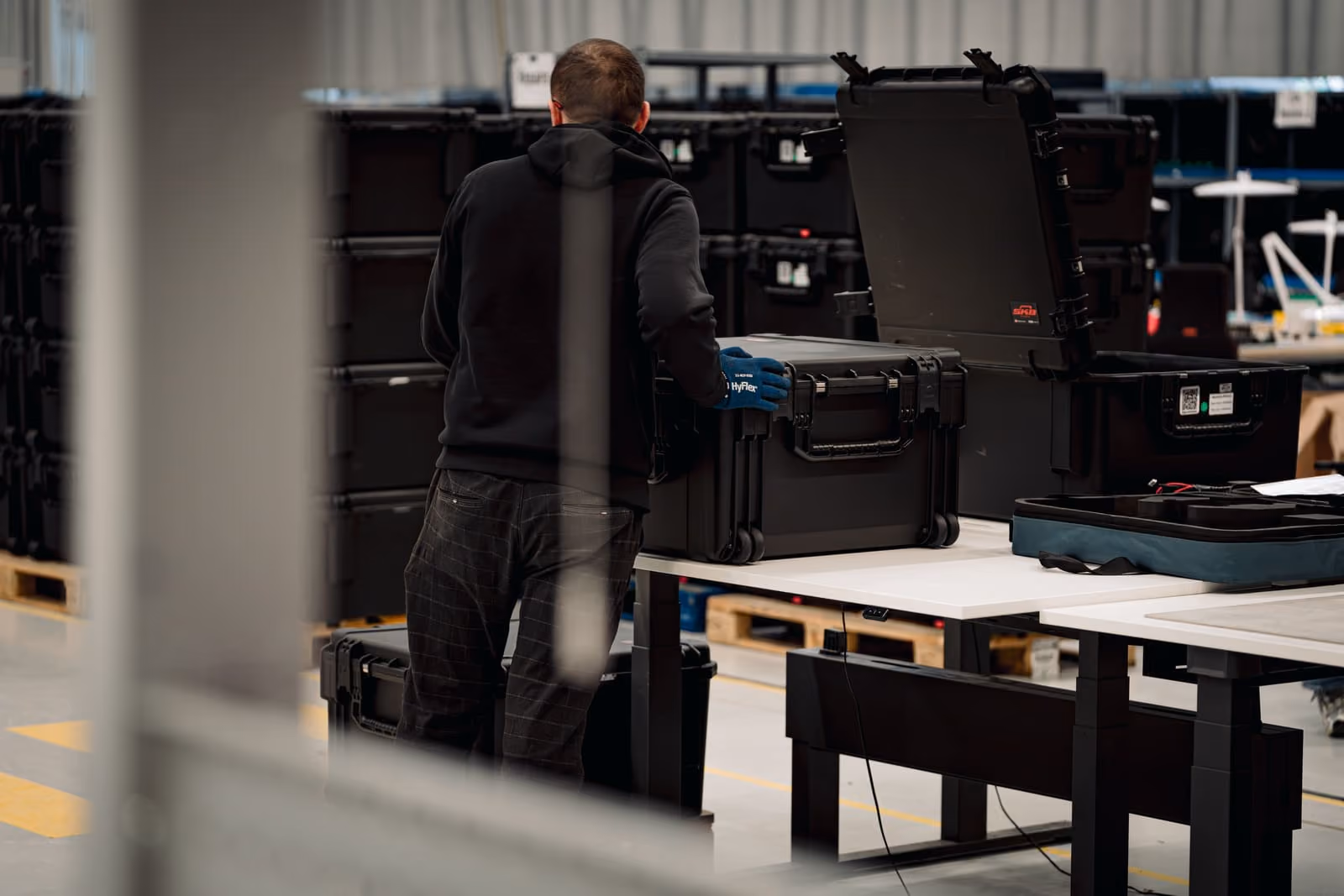 Worker in gloves handling large black equipment cases stacked in an industrial warehouse.