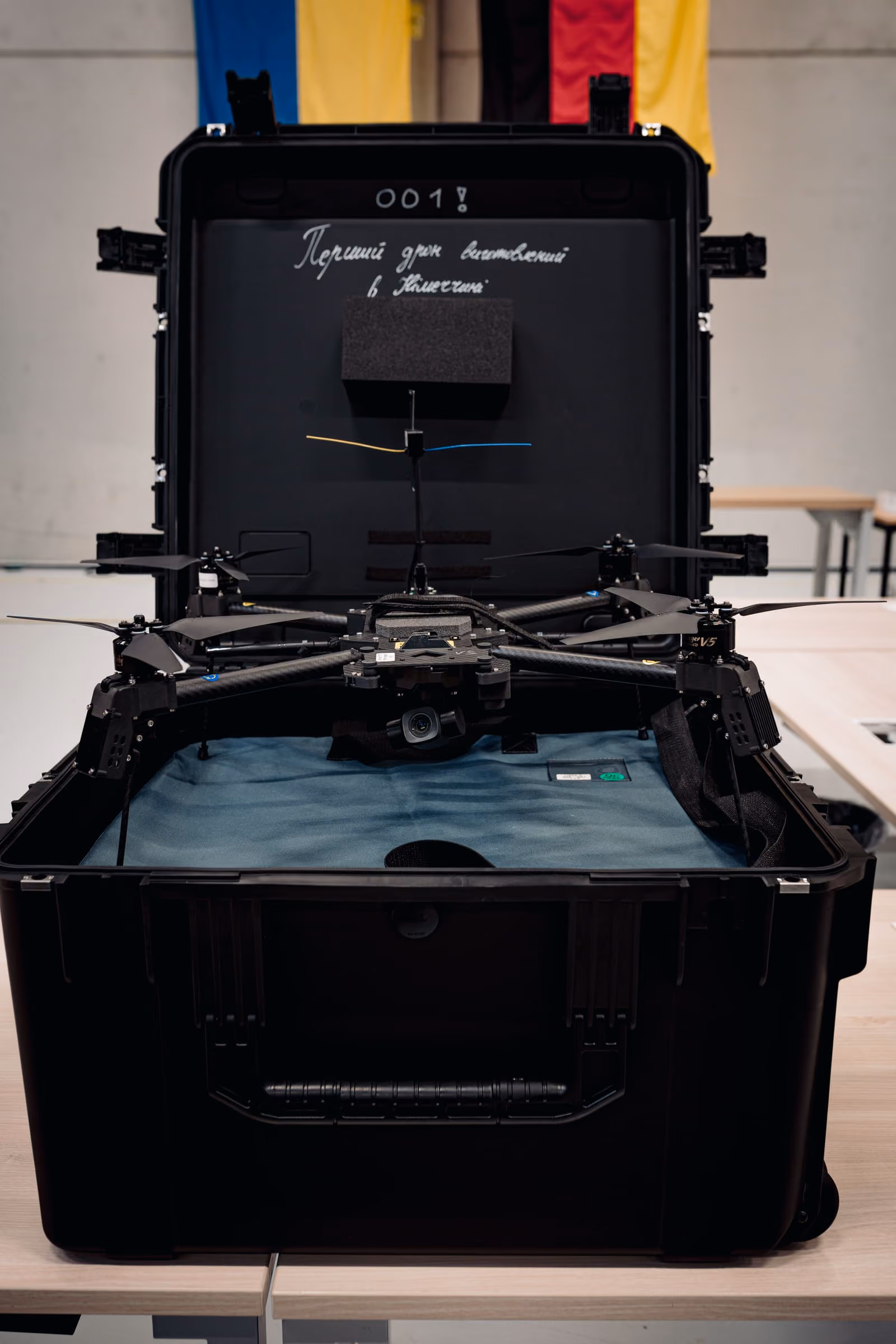 Black drone with four propellers inside an open black carrying case on a table, with colored wires and flags behind.