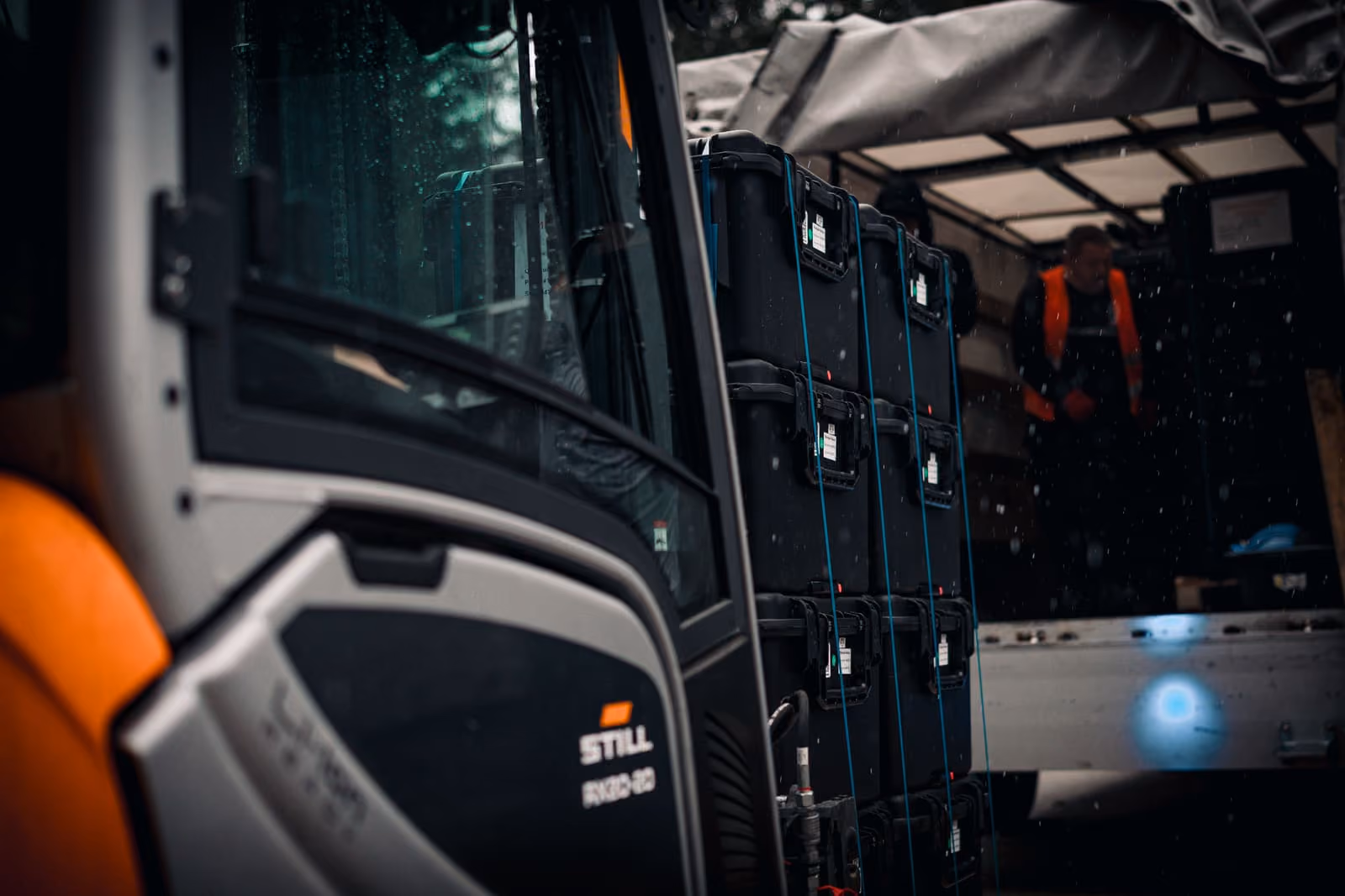 Forklift moving stacked black storage cases while worker stands in truck wearing orange safety vest.