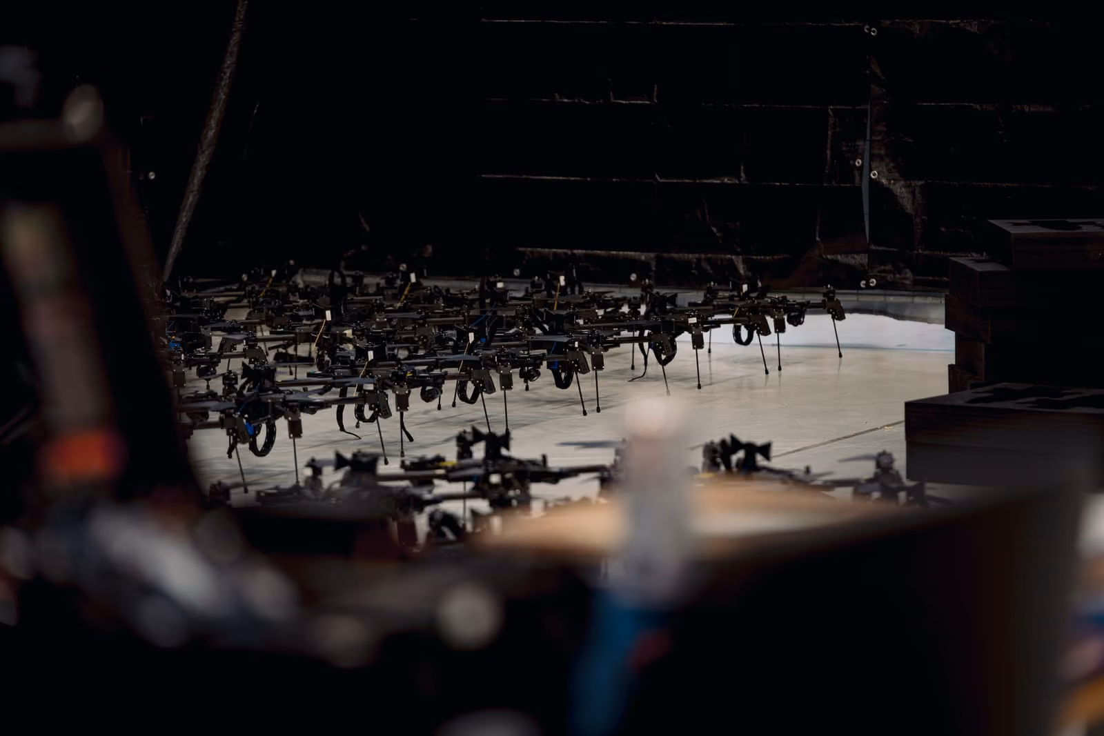 Numerous small Quantum Frontline Industries drones with rotary wings assembled on a floor against a dark brick wall backdrop.