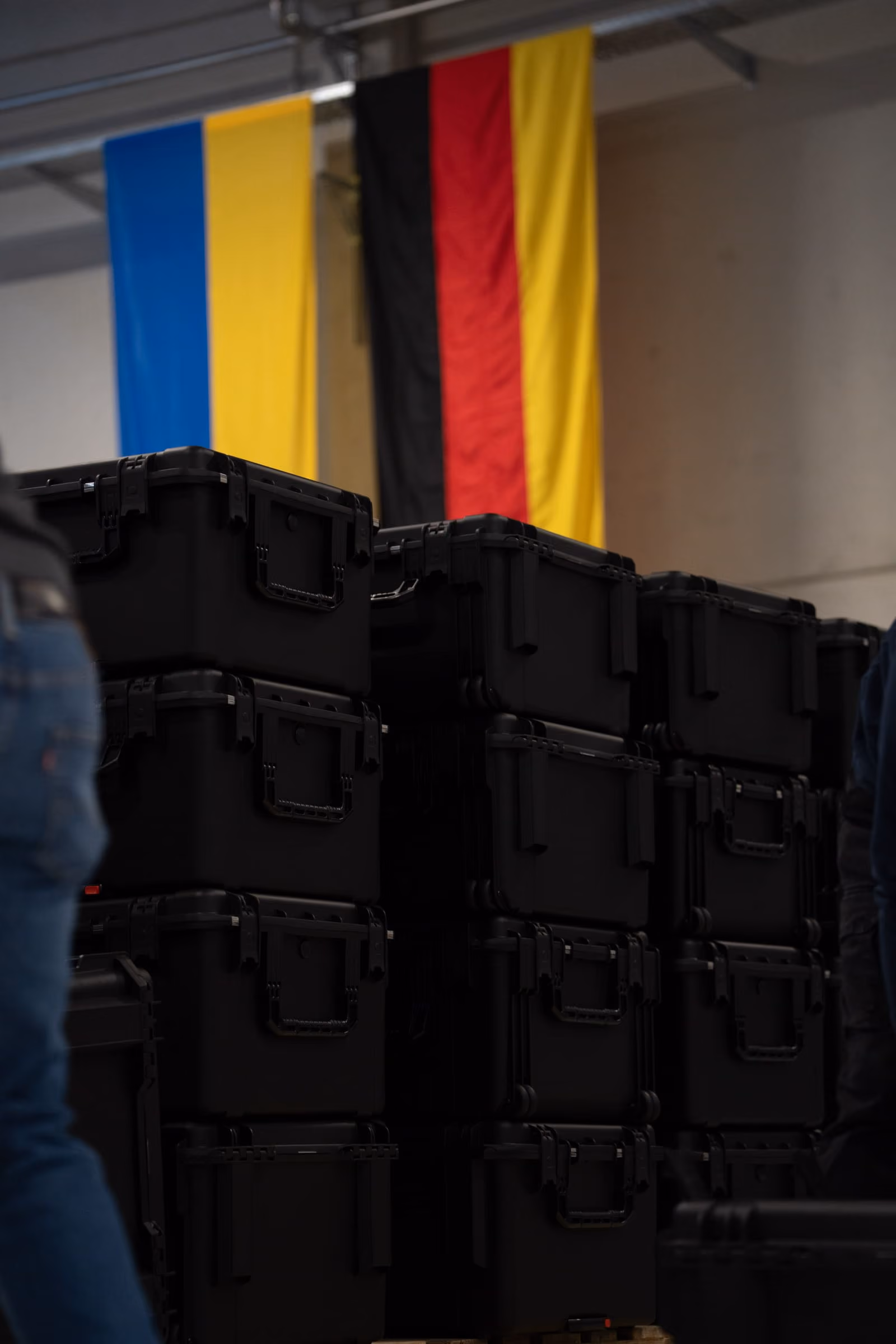 Stacked black Quantum Frontline Industries cases on pallets with blurred Ukrainian and German flags in the background.