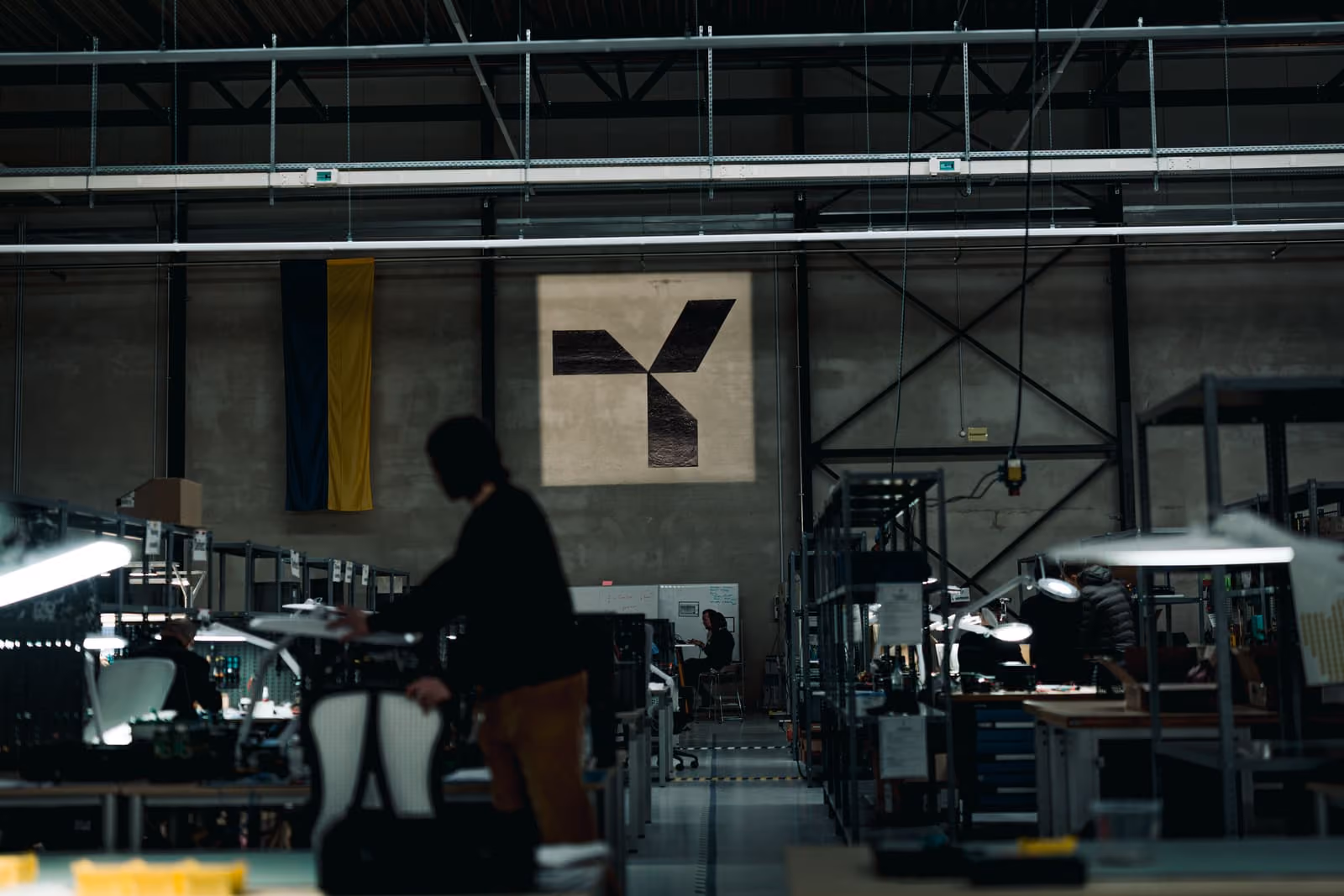 Dimly lit industrial workspace with people working at desks and a large logo on the wall.
