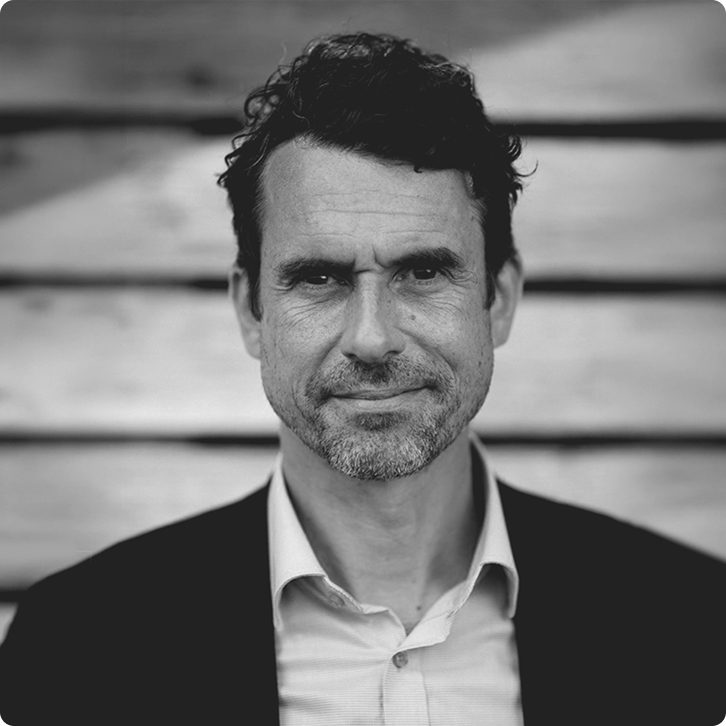 Black and white portrait of Jonas with short curly hair and stubble, wearing a collared shirt and jacket.