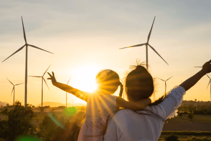 Silhouette of a woman holding a child with arms raised, standing near wind turbines at sunset.