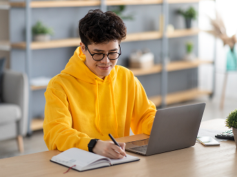 Person wearing a yellow hoodie and glasses working on a laptop and writing in a notebook at a desk.