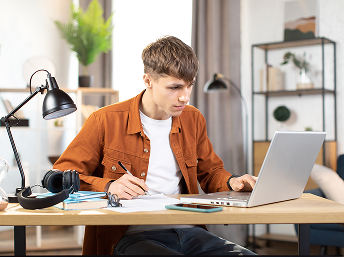 Young man in an orange jacket working on a laptop with a notebook and pen at a desk in a modern room.