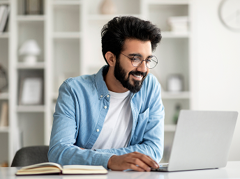 Man with glasses and beard smiling while working on a laptop at a desk with books.