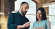 Man and woman standing indoors, looking at a tablet and smiling.