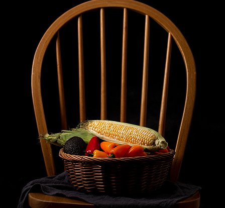 Wicker basket with corn, avocado, and assorted colorful peppers on a wooden chair with vertical slats against a black background.