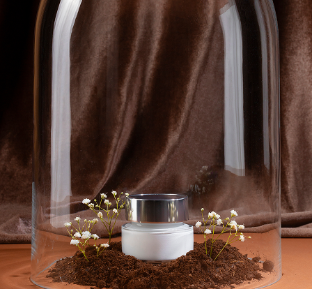 A white cosmetic jar with a silver lid placed on soil, surrounded by small white flowers, all under a glass dome with a brown fabric backdrop.