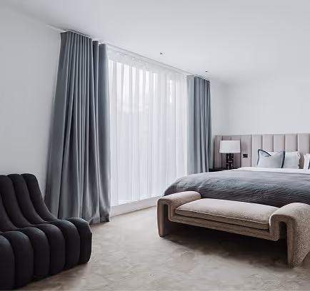 Modern bedroom with gray bed, upholstered bench, black chair, sheer curtains, and soft lighting.