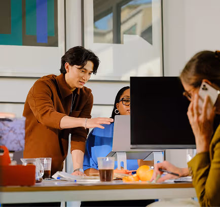 Two people collaborating at a desk with a laptop and monitor while another talks on phone nearby.