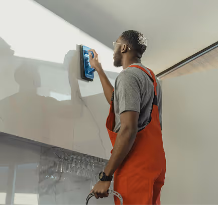Man in red overalls cleaning a high wall with a blue cloth indoors near hanging glasses.