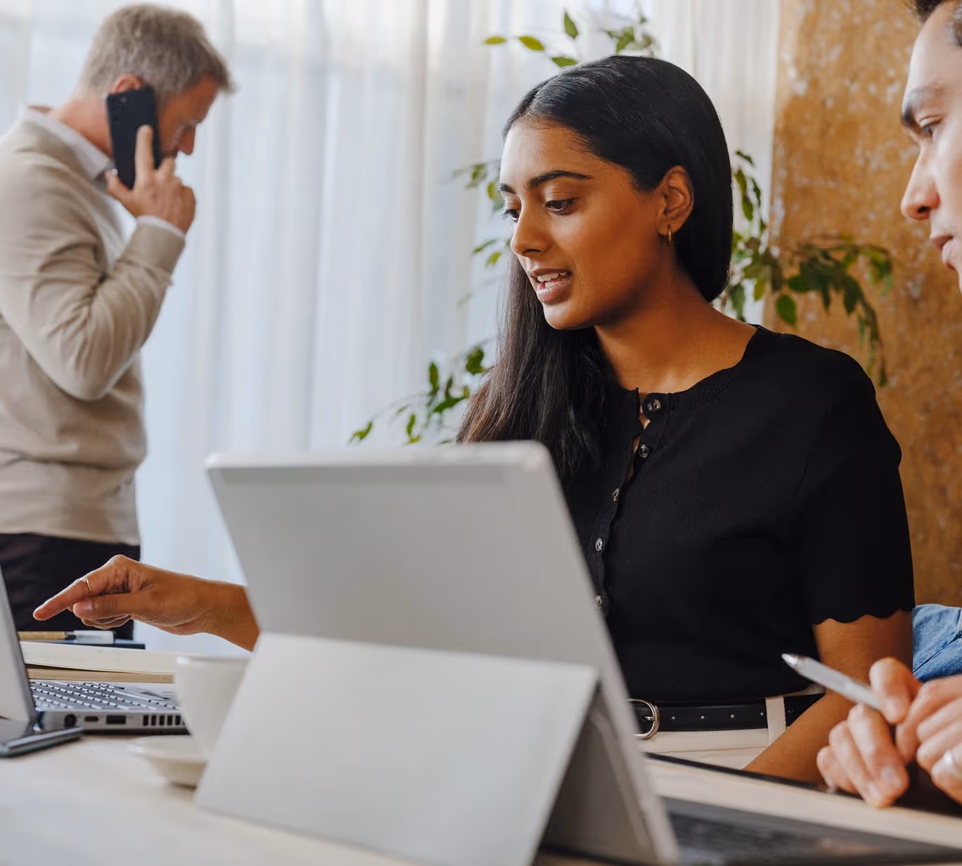 Woman pointing at a laptop screen while a man beside her writes and another man talks on phone in background.