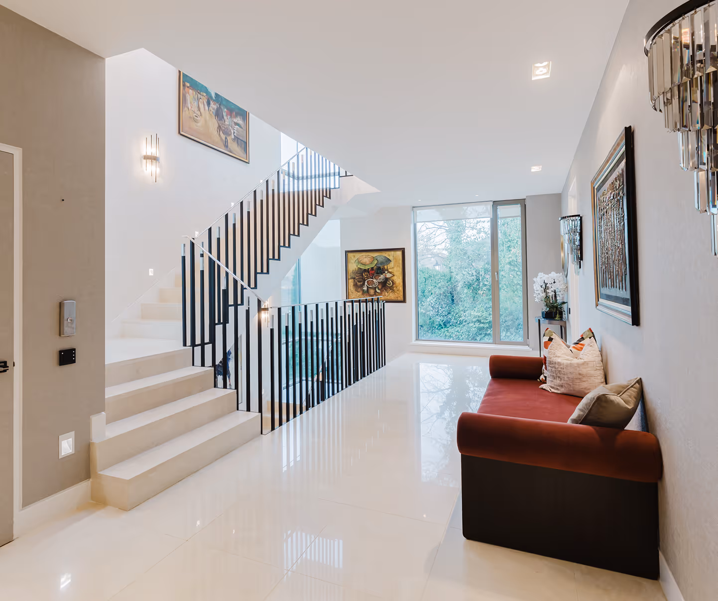 Modern hallway with white stairs, a red sofa with pillows, large window, and artworks on walls.