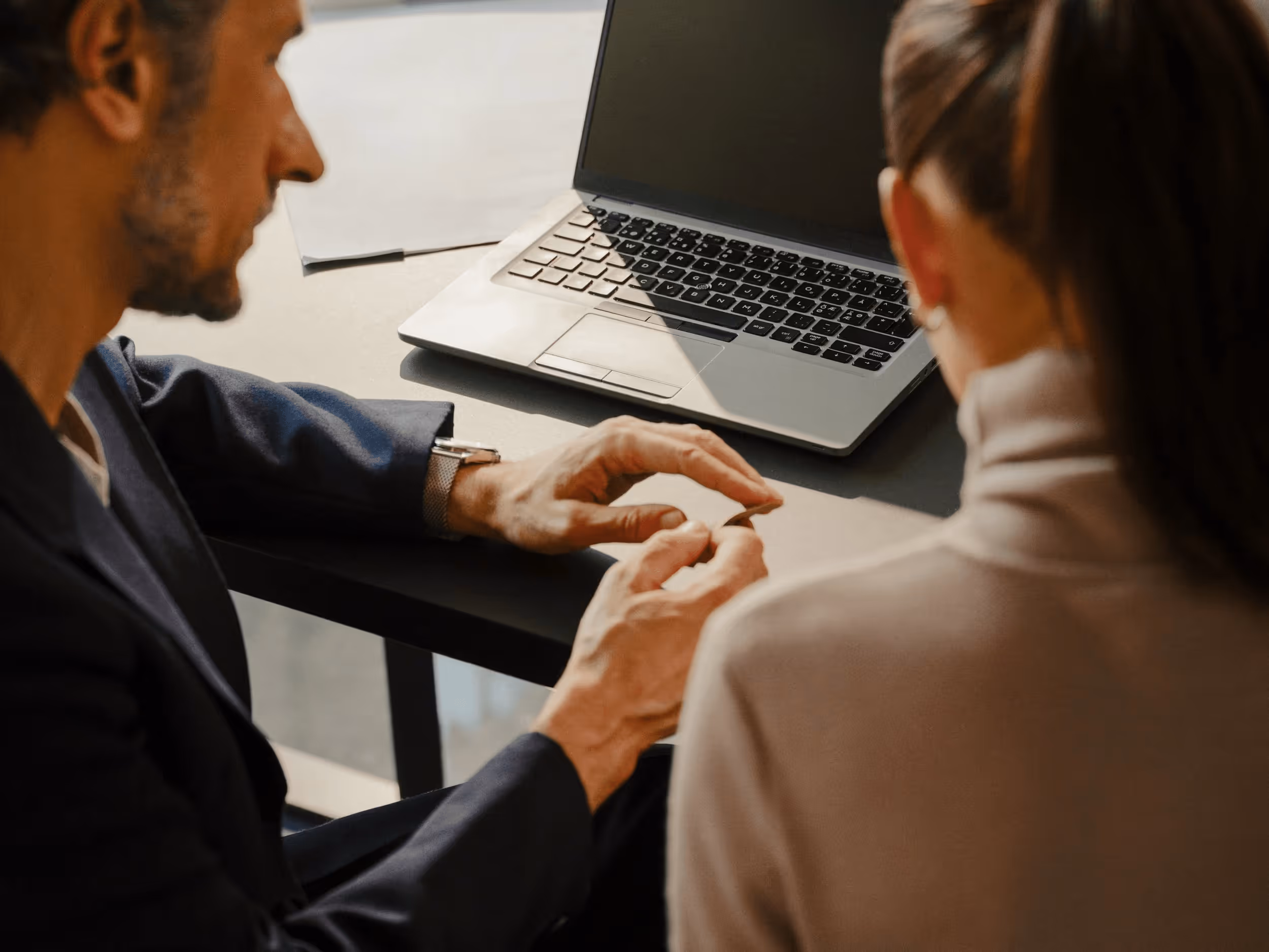 Two people seated at a table, one gesturing with hands near a laptop with a black screen.