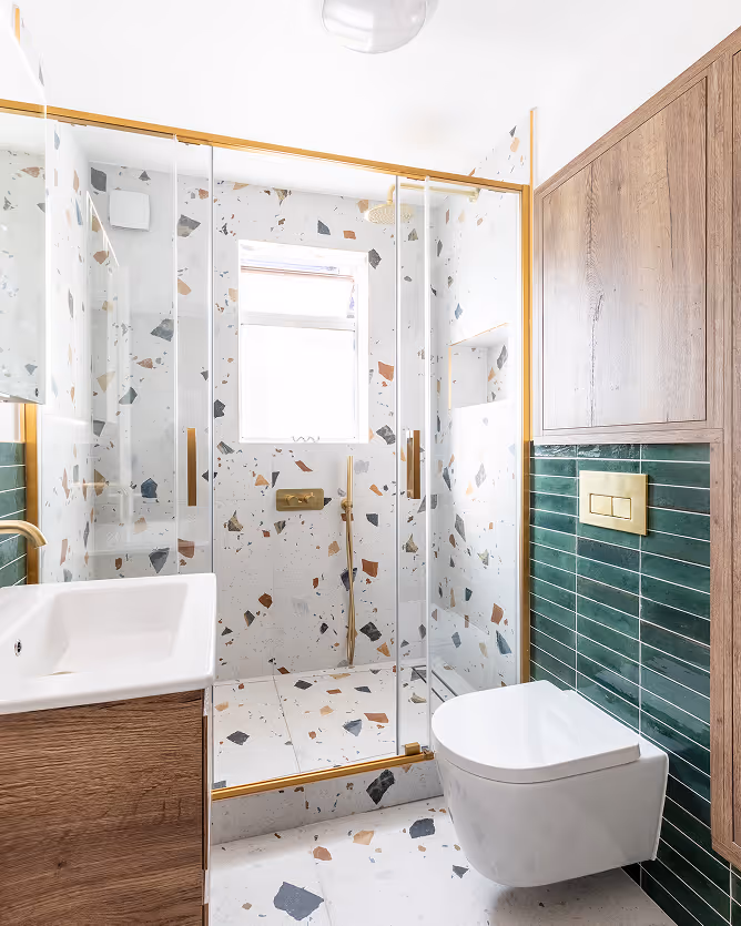 Modern bathroom with terrazzo tiles, glass shower enclosure with gold trim, white wall-mounted toilet, and wood cabinetry.