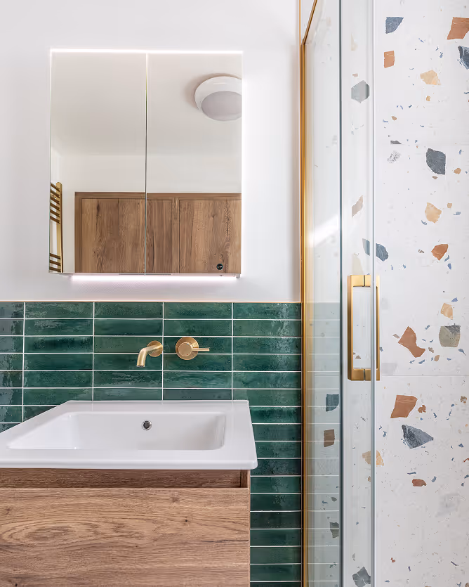 Bathroom with white sink, green tiled wall, gold faucet, wooden cabinet, glass door, and mirror cabinet.