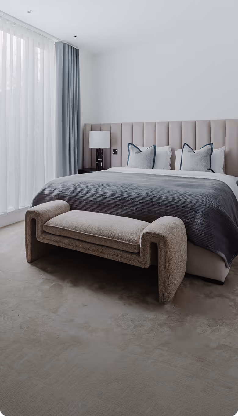 Minimalist bedroom with a bed, gray blanket, decorative pillows, beige bench, and floor-to-ceiling curtains.
