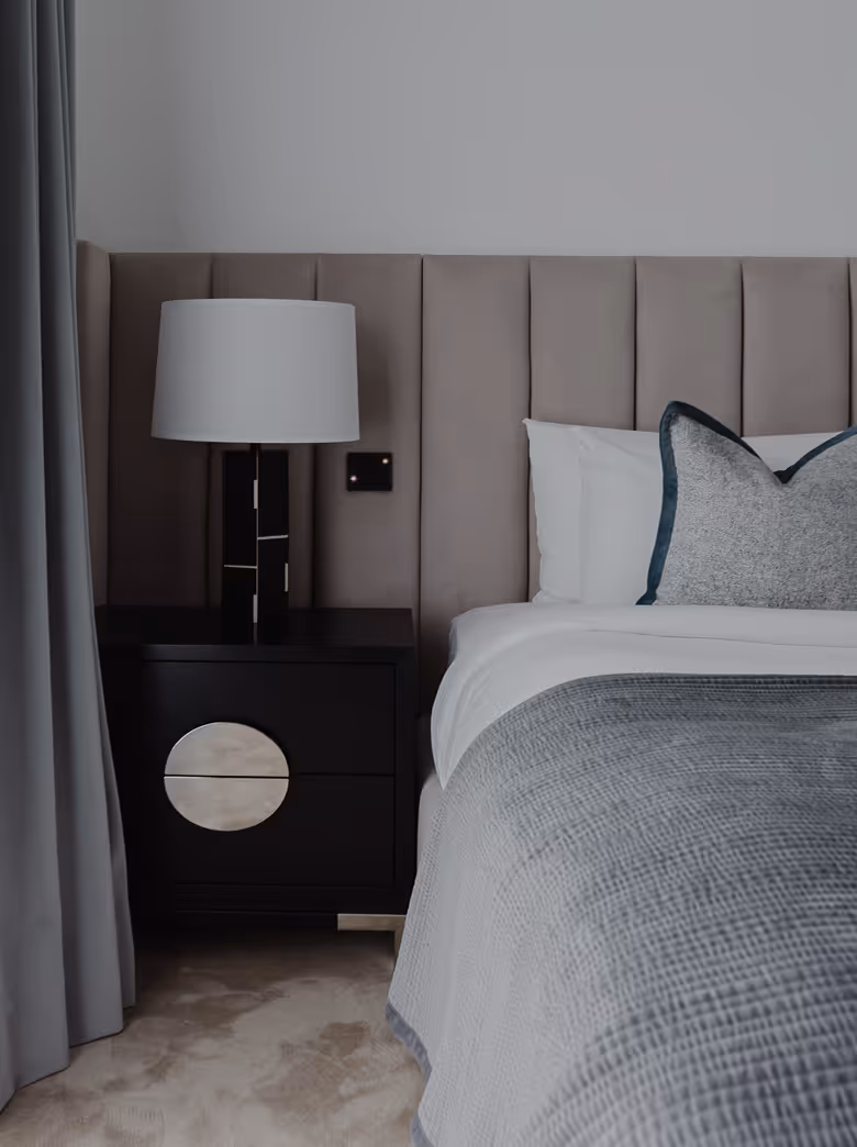 Modern bedroom corner with upholstered headboard, black nightstand, lamp, grey bedding, and curtains.
