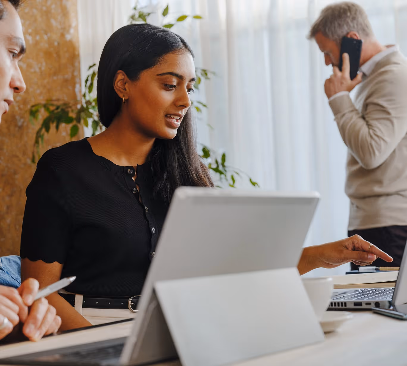 Woman pointing at a laptop screen with a man on phone in the background in an office setting.