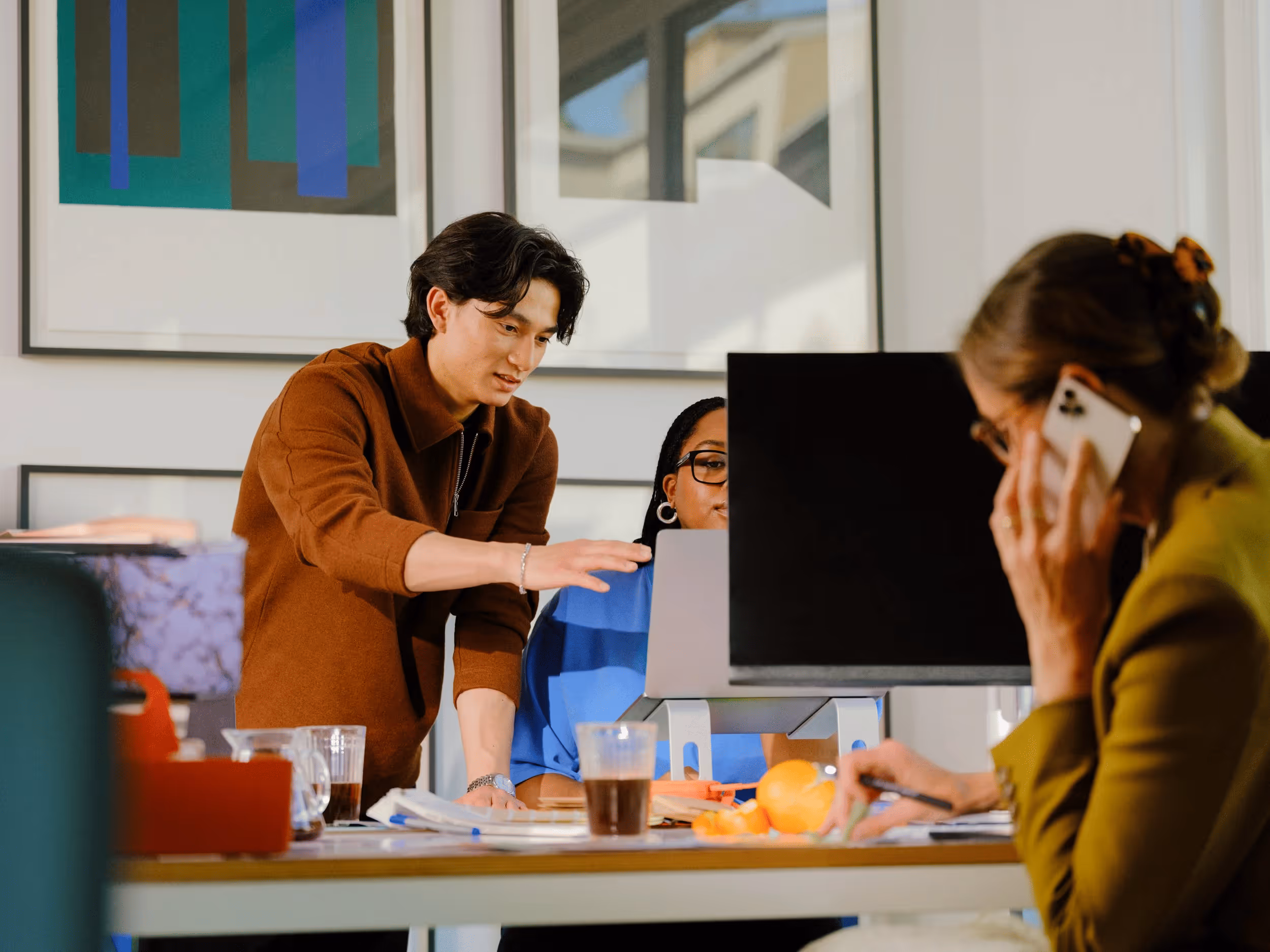 Two coworkers collaborate at a desk with computers while another talks on phone nearby.