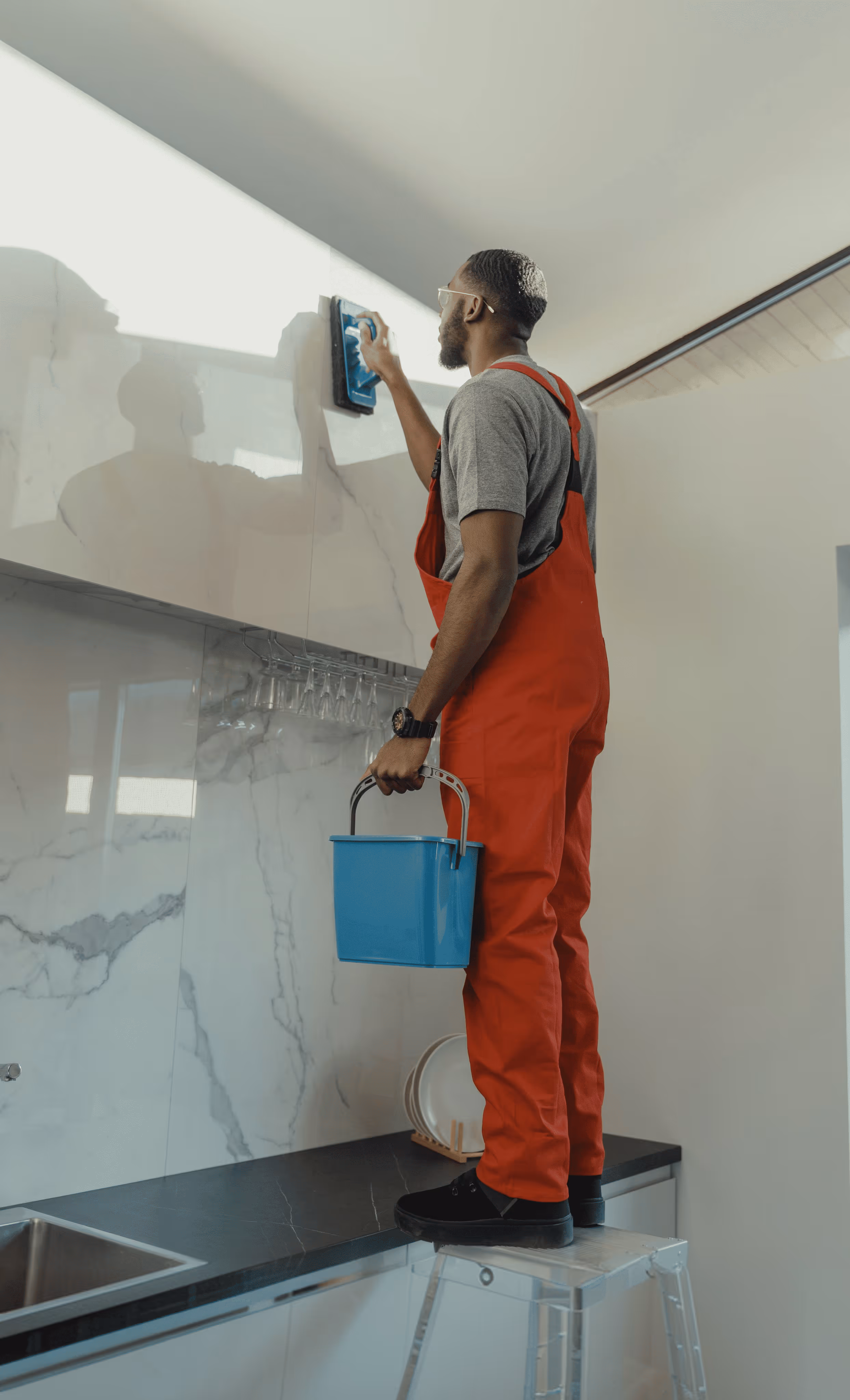 Man in red overalls standing on a stepladder cleaning a kitchen cabinet with a blue bucket in hand.
