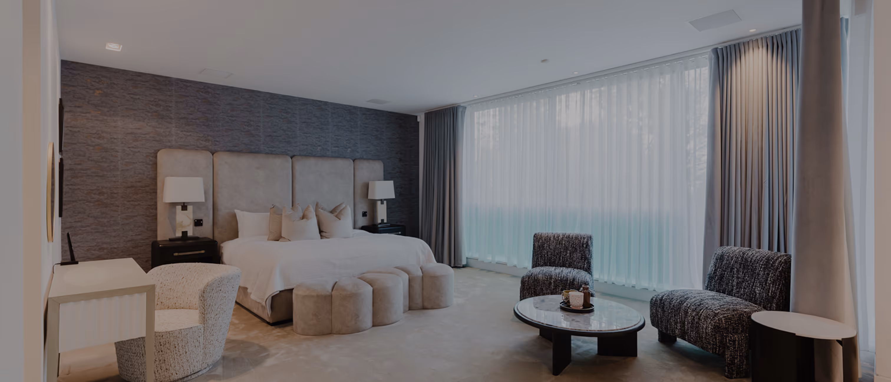 Modern bedroom with large bed, beige decor, round ottomans, two chairs, and floor-to-ceiling window with curtains.