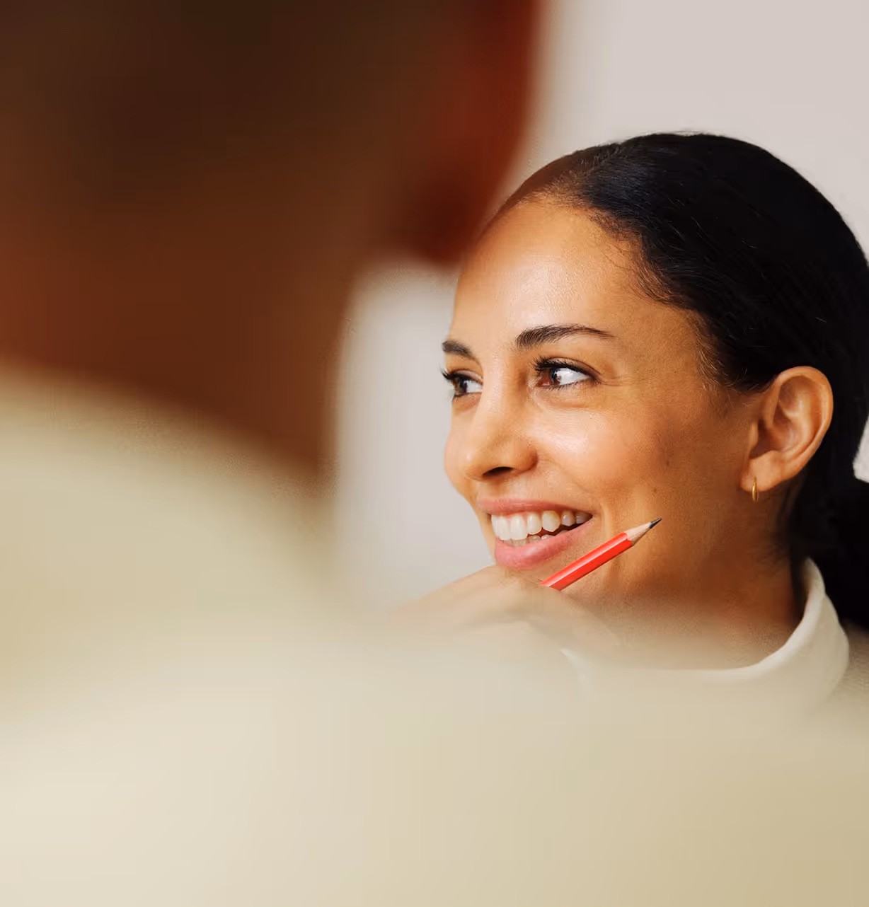 Smiling woman holding a red pencil near her chin looking to someone blurred in foreground.