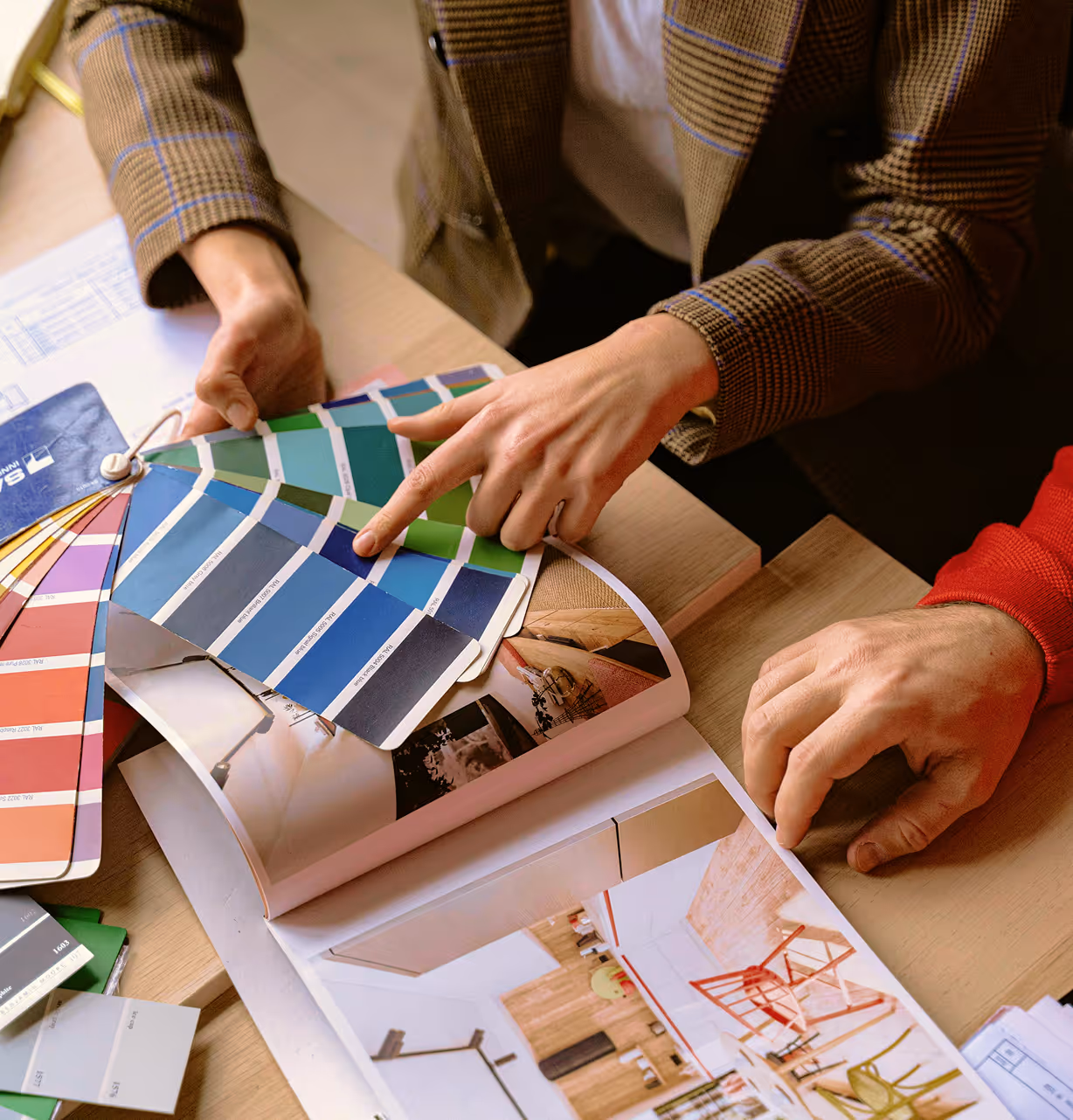 Two people comparing a blue-green color swatch with an interior design magazine on a wooden table.