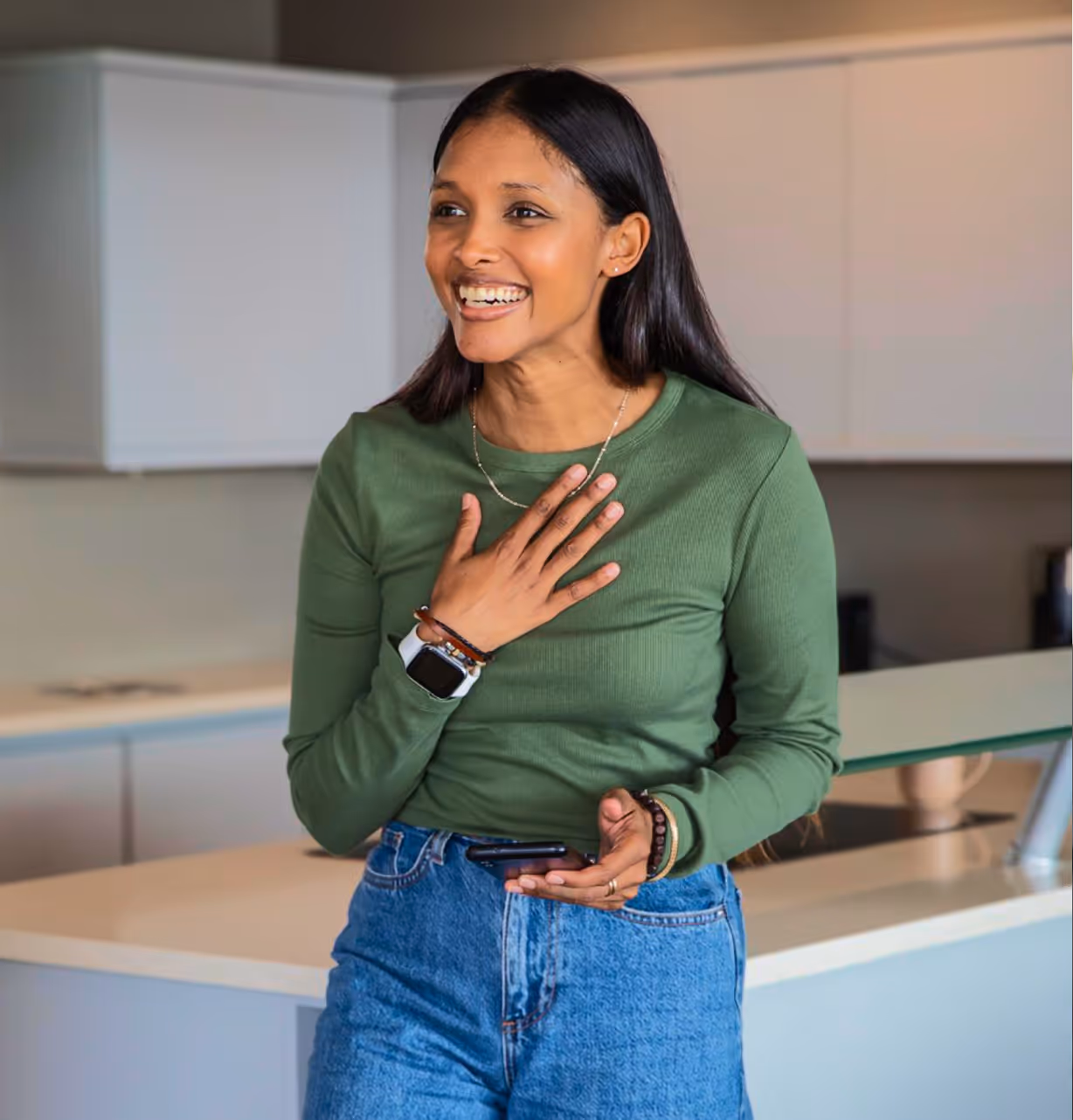 Smiling woman in green shirt and jeans holds phone and touches chest in modern kitchen.