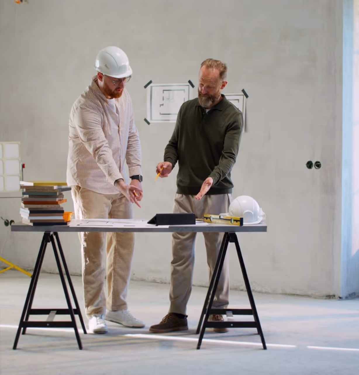 Two men reviewing architectural plans on a table in an unfinished room with construction gear nearby.