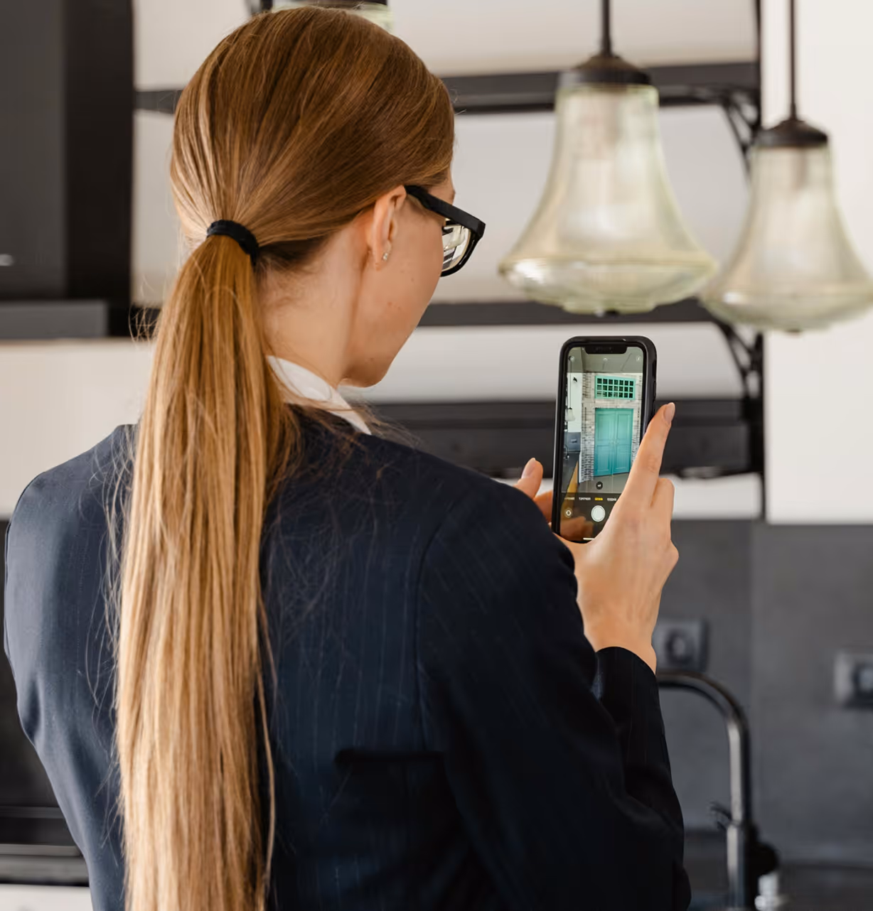 Woman with long hair in a ponytail takes photo of green door using a smartphone indoors.