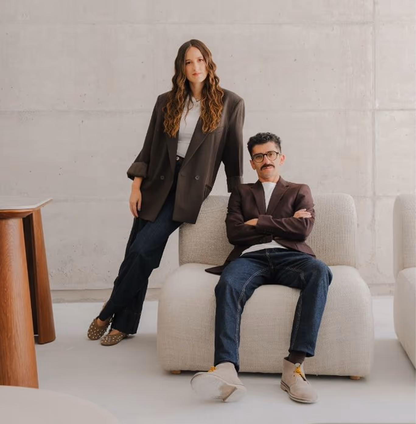Man sitting on a light beige sofa with arms crossed and woman standing beside it leaning, both dressed in blazers.