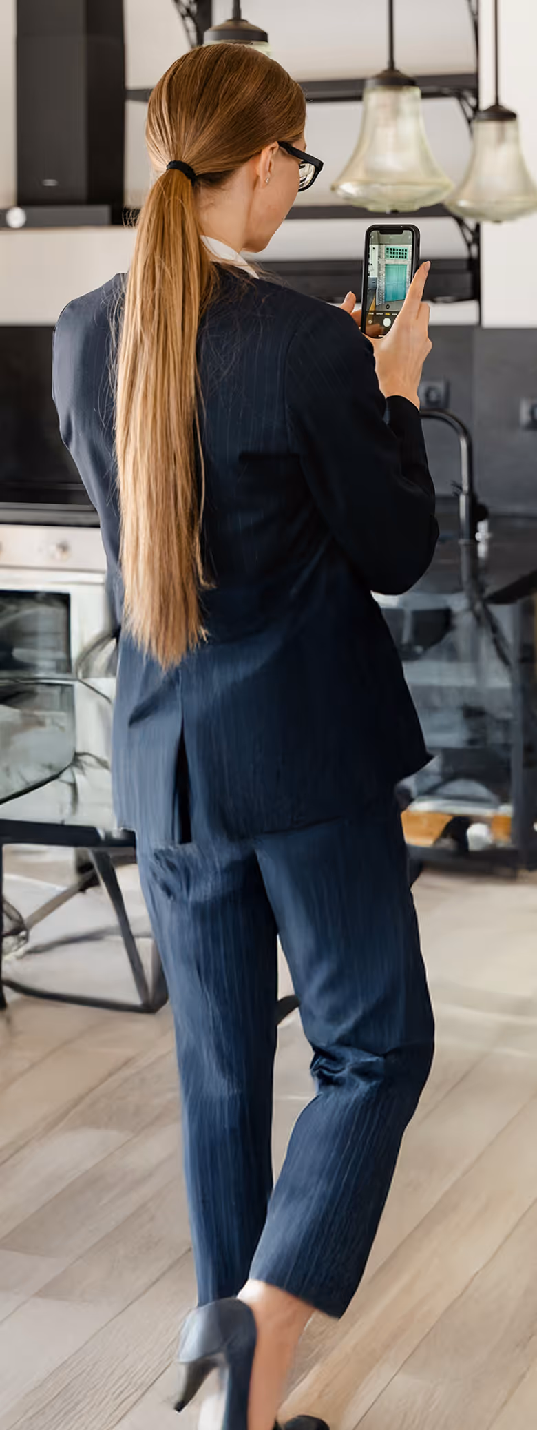 Woman in a dark suit with long hair tied back, taking a photo with a smartphone in a modern kitchen.