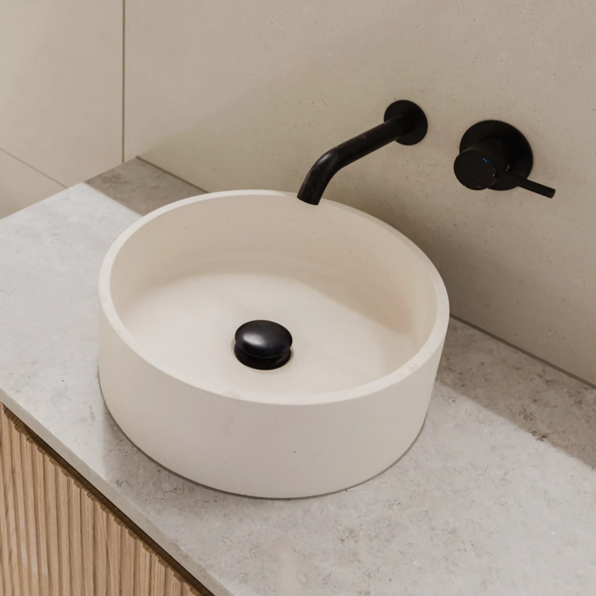 Modern round white bathroom sink with a black stopper on a gray stone countertop, featuring a matte black wall-mounted faucet and handle against a beige tiled wall.