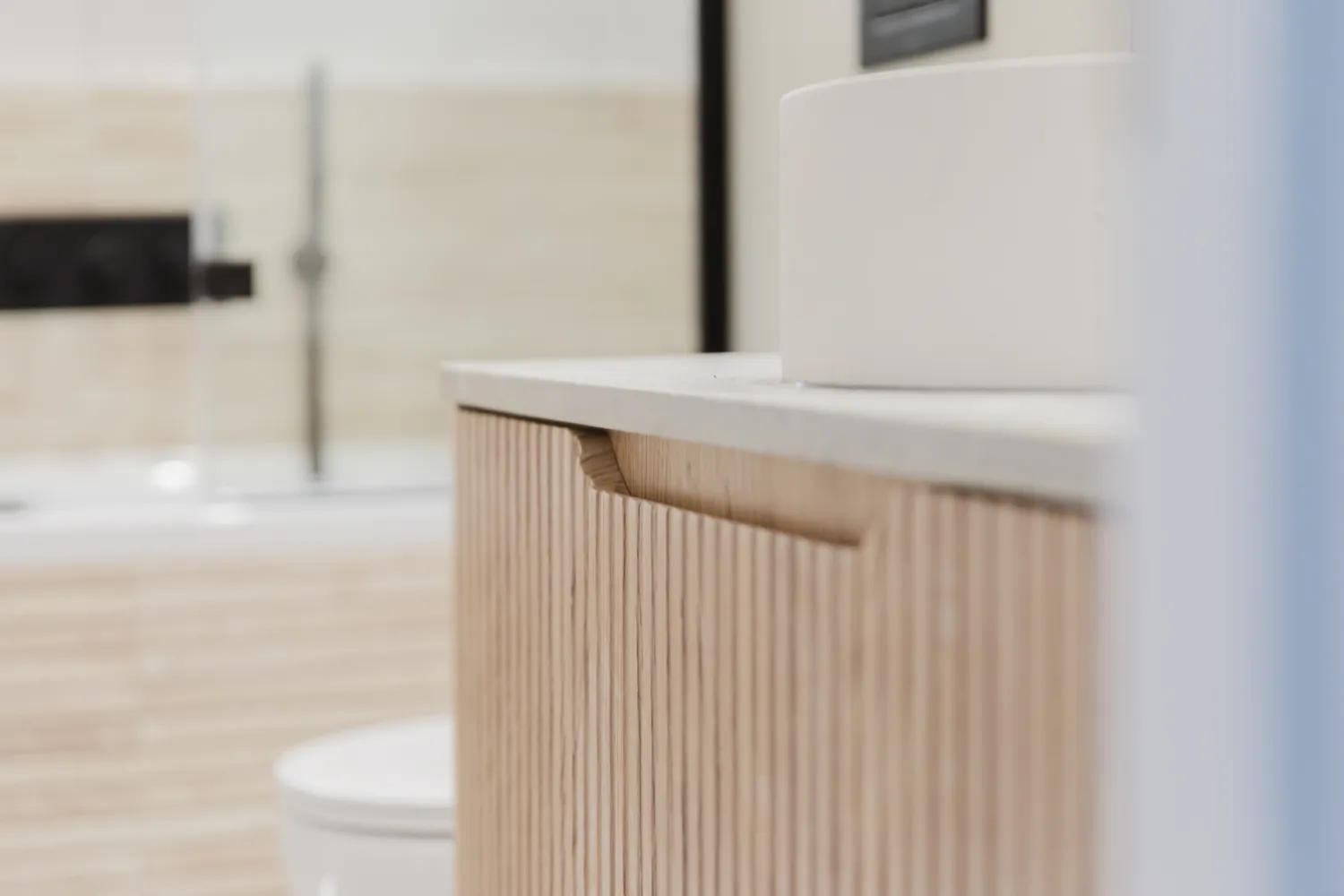 Close-up of a bathroom vanity with vertical wood slat cabinet doors and a white countertop, with a blurred toilet and bath fixtures in the background.