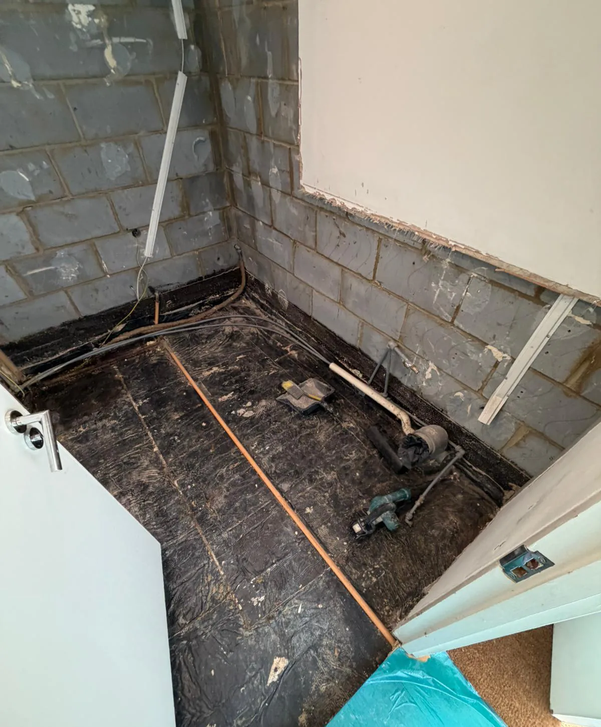 Small room under renovation with exposed gray cinder block walls, a concrete floor covered by black protective sheeting, plumbing pipes along the edges, scattered construction tools, and an open white door.