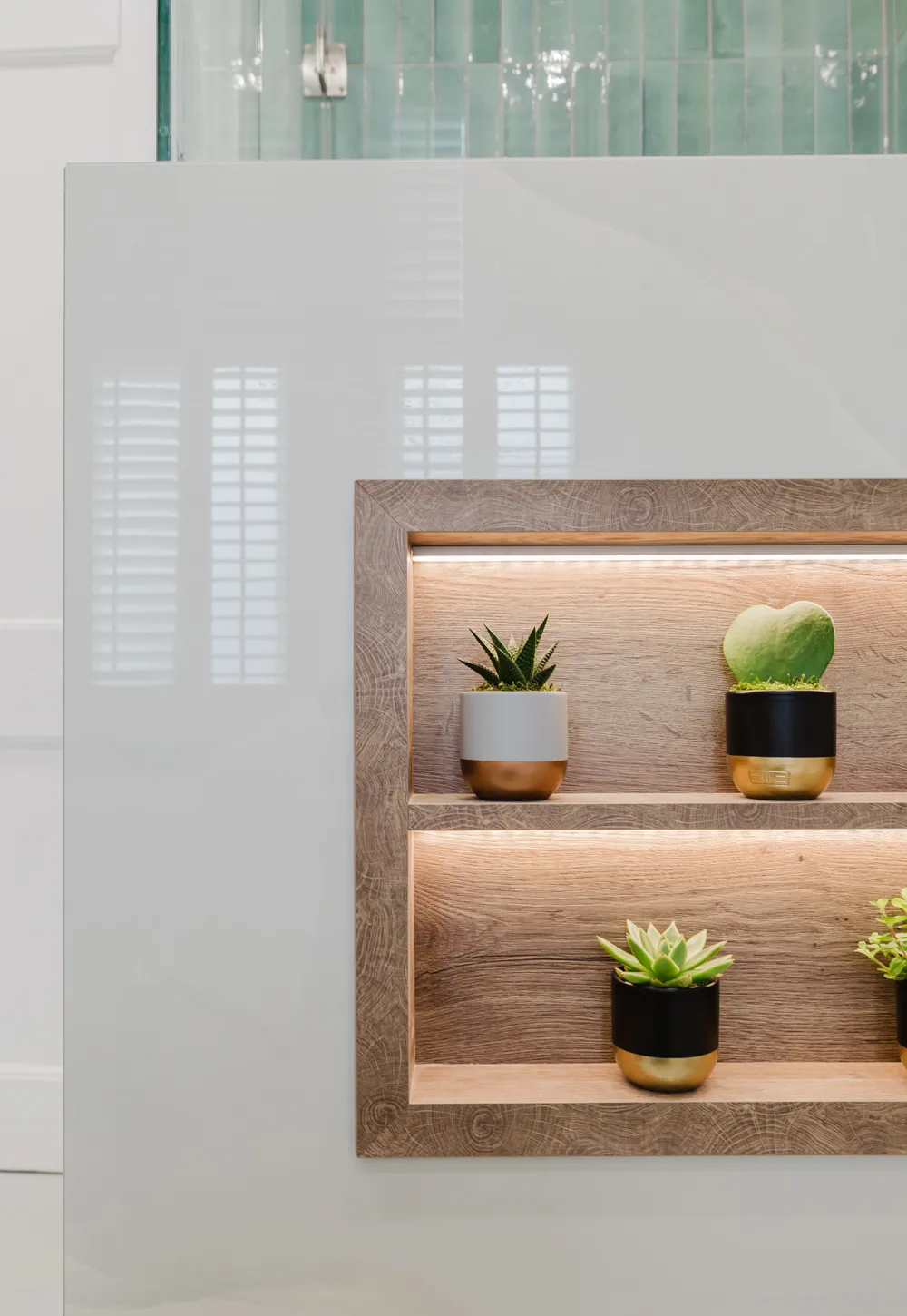 A wooden recessed shelf with two levels, each lit by soft lighting, holding four small potted succulent plants in stylish black and white pots with gold bases.