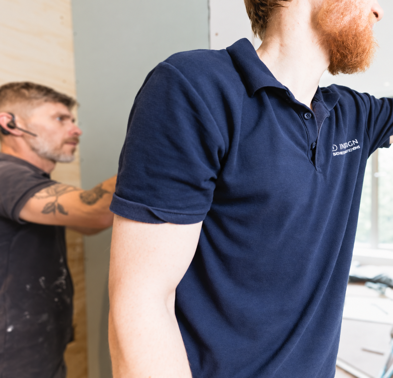 Two men working indoors on a wall; the man in the foreground has a red beard and wears a navy blue polo shirt, while the man in the background has a tattoo on his arm, wears a headset, and focuses intently on the task.
