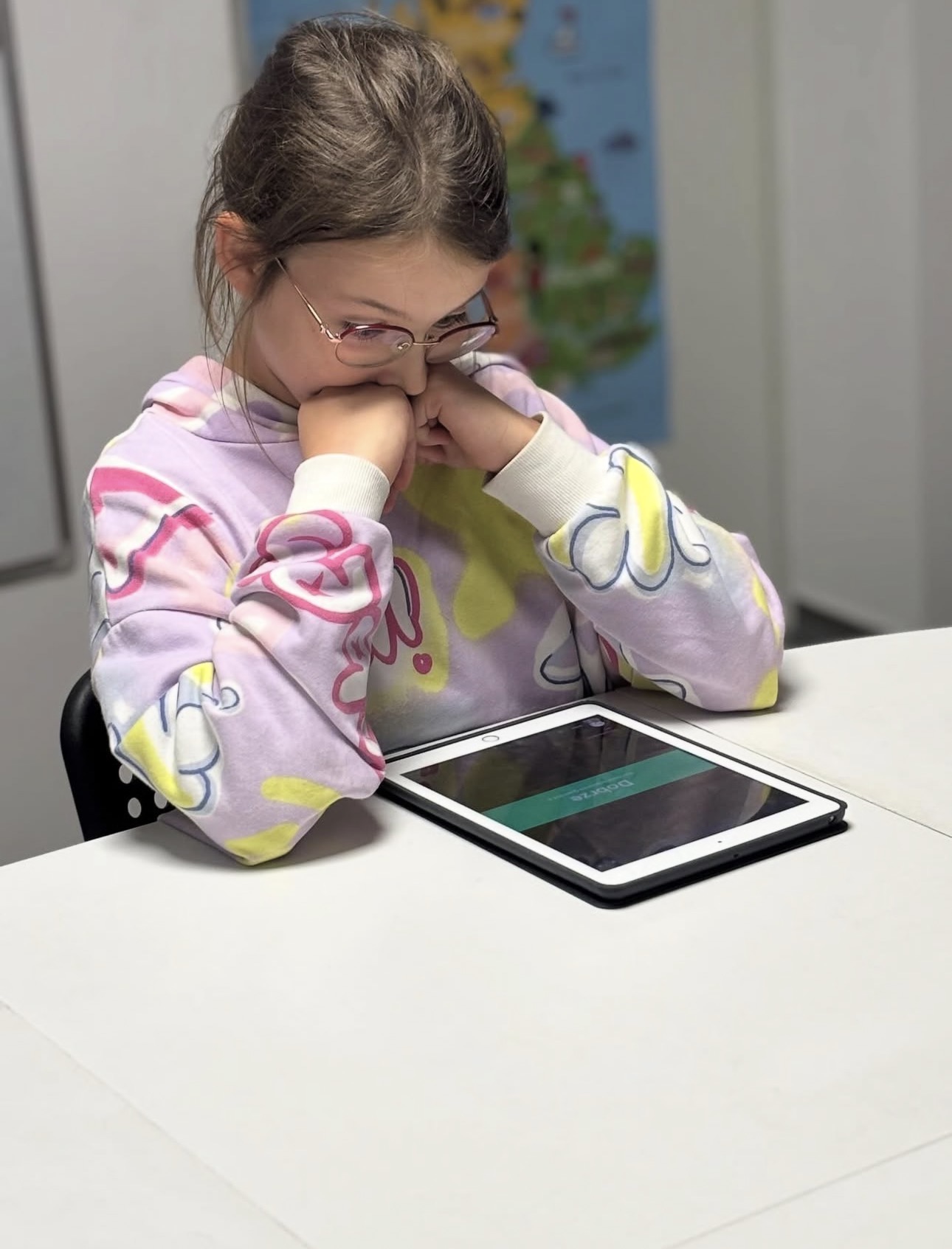 Girl with glasses in colorful hoodie looking at tablet on white table in a classroom.
