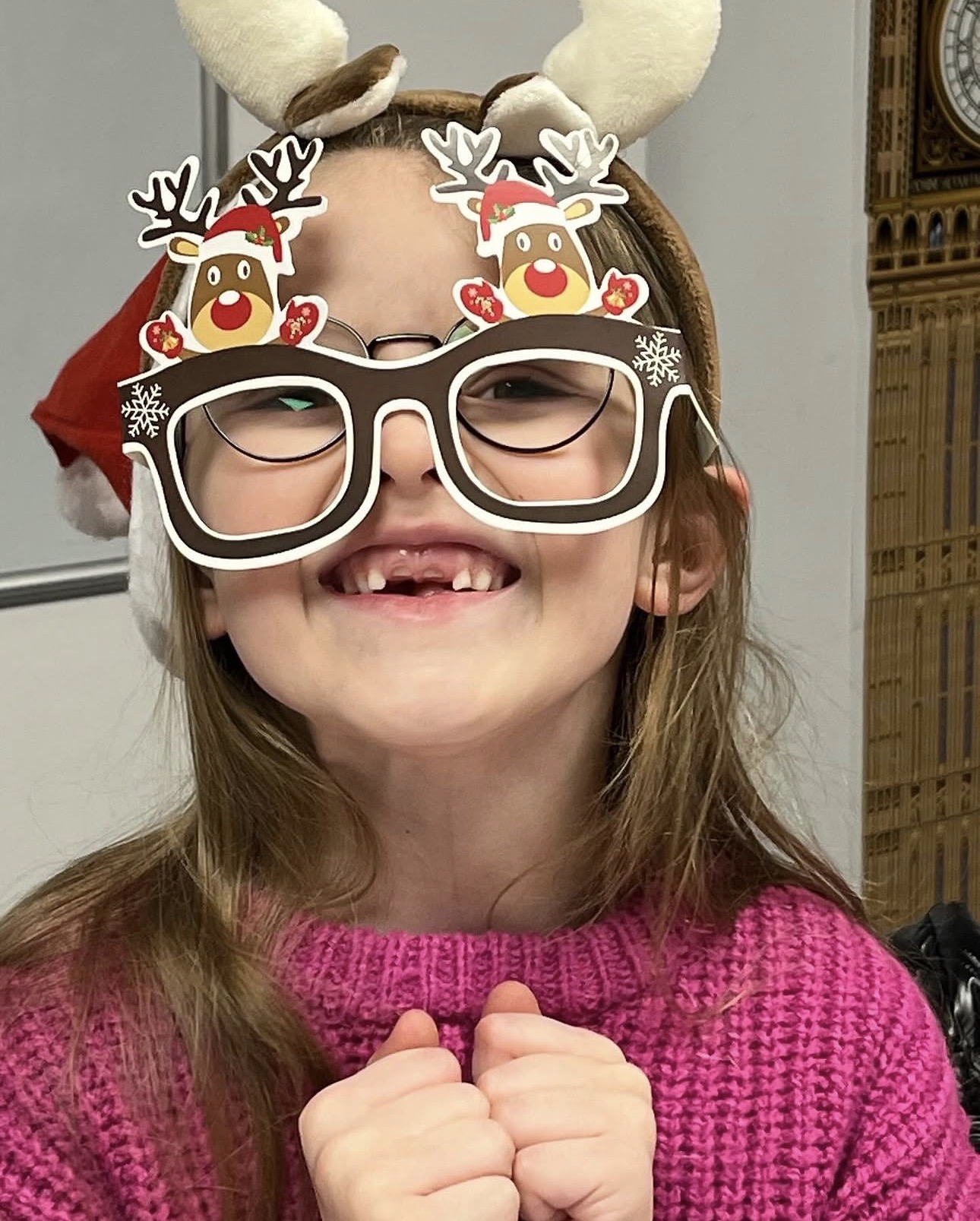 Smiling child wearing reindeer glasses with Santa hats and a pink sweater, showing missing front teeth.